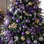 Purple Christmas tree decorated with amethyst ornaments, gold and silver accents, crystal ornaments, metallic ribbons, beaded garlands, plum berry sprigs, and a dramatic topper, glowing under warm lights with a blurred fireplace and frosted window behind.