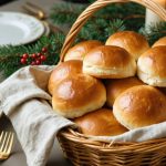 Linen-lined basket filled with golden, fluffy vegan dinner rolls brushed with plant-based butter, on a festive holiday table with softly blurred plates, greenery, and candlelight.