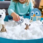 Eye-level close-up of a child’s hands playing in a cotton “snow” sensory bin with polar bear, arctic fox, snowy owl, and penguin figurines, with handmade paper plate polar bear, paper bag penguin puppet, and handprint reindeer ornament nearby; soft daylight and blurred twinkle lights in the background.