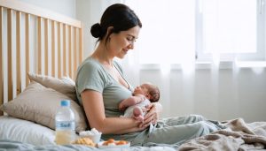 Postpartum mother in loose loungewear sitting on a bed, cradling her newborn with a small pillow against her abdomen, soft daylight from a window, bedside table with water and snacks, and a blurred crib and textiles in the background.