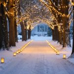 Snow-dusted driveway at dusk illuminated by warm LED path lights and flickering luminarias, with string lights hanging between trees and subtle entrance spotlights; house and evergreens softly blurred in the background.