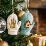 Close-up of personalized Christmas ornaments on a tree—a clear glass bauble with a tiny gold handprint, a carved wooden snowflake, a felted mitten with a pet portrait, and a hand-painted ceramic house—lit warmly with blurred fireplace and wrapped gifts in the background.