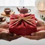 Two pairs of hands exchange a red furoshiki-wrapped present tied with a gold ribbon at eye level in soft daylight, with a blurred background of a tea set, unbranded chocolates, paper and geometric lanterns, and winter greenery.