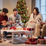 Multigenerational family—toddler, teen, parents, and grandparent—exchanging personalized gifts beside a decorated Christmas tree, warm golden lighting, cozy living room with fireplace and snowy window in the background.