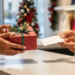 Close-up of hands exchanging a small wrapped present and a plain gift receipt across a retail returns counter with softly blurred holiday decor and shopping bags in the background, no readable text or logos visible.