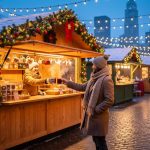 Eye-level view of a festive American Christmas market at dusk, with a wooden chalet stall decorated in evergreen garlands as a vendor hands a steaming cup of mulled wine to a visitor on a cobblestone walkway, warm string lights, light snowfall, and a softly blurred city skyline in the background.