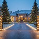 Driveway entrance at blue-hour dusk with light snowfall, permanent LED track lights lining both sides, stone pillars wrapped in rope lights, and mature evergreen trees wrapped in warm white strands with subtle red and green accents; house and landscaped shrubs softly visible in background.