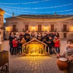 Spanish plaza at twilight with families singing around a warmly lit nativity, a steaming chestnut vendor in the foreground, a child holding a zambomba drum, and silhouettes of the Three Wise Men parade approaching under golden string lights and historic stone buildings.