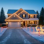 Eye-level view from a driveway entrance showing warm white icicle lights on a suburban roofline, red-and-white candy cane lights lining the driveway, and illuminated reindeer on a snow-dusted lawn at twilight, with softly blurred neighboring houses and evergreens in the background.