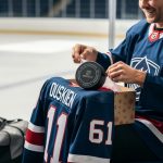 Hockey fan unwrapping a personalized ice hockey gift with a custom jersey and a commemorative hockey puck visible on the box, with a blurred rink background.